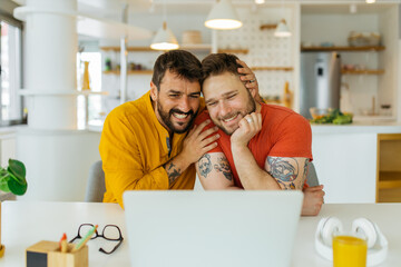 A happy, gay couple sits together at the dining table and having a video call with their friends.