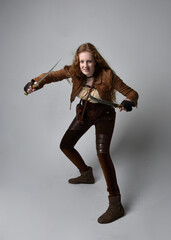 Full length  portrait of beautiful young woman with long red hair, wearing steampunk inspired costume knife weapons isolated on studio background.