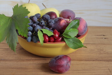 There are grapes, dogwood, plums and apples in a clay bowl.