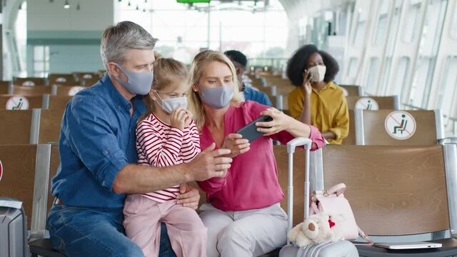 Young caucasian spouses wearing protective masks sitting in the airport and taking selfie photos with their daughter to the camera of smartphone