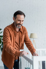 Positive man standing near blurred baby crib at home