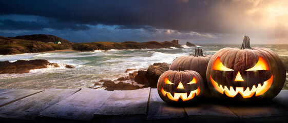 Sunset at the coast with the spooky evil glowing eyes of Jack O' Lanterns on the right of a wooden bench on halloween night.