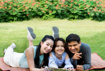 Happy family resting together on the grass having fun outdoor. Mother, father, daughter laying on mat. Picnic in the park. Beauty nature scene with family outdoor lifestyle. Happiness in family life.