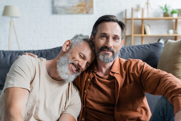 Mature man hugging smiling homosexual partner at home - obrazy, fototapety, plakaty