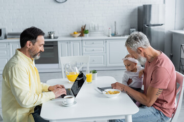 Homosexual man using laptop near daughter and partner with laptop in kitchen