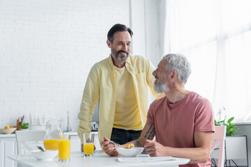 Smiling homosexual man standing near partner and breakfast in kitchen