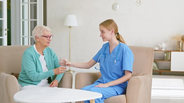 Young doctor gives water to senior woman at table in room
