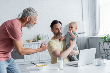 Same sex couple with baby bottle and child near laptop in kitchen
