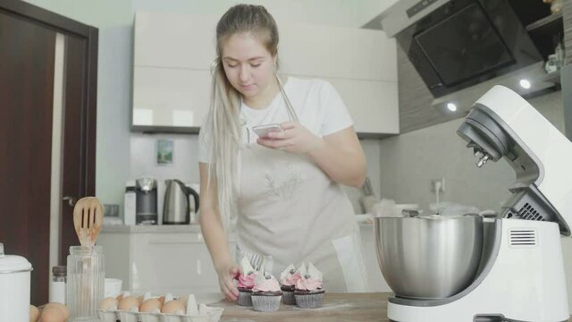 A beautiful girl in the kitchen takes pictures of prepared cupcakes on her phone. Handmade work