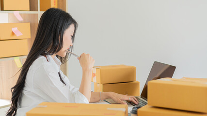 Startup, small business, SME, owner of Asian female entrepreneurs working with boxes. Smile and check the sales deficit using a computer. After she succeeded in thinking differently in online SME busi