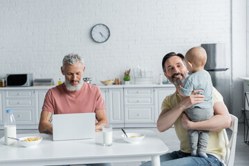 Obraz premium Smiling homosexual man holding baby girl near partner using laptop and breakfast in kitchen
