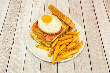 Double-decker sandwich with fried egg with yolk for dipping, grilled bread with butter, sliced cheese, fried bacon and side of French fries with lettuce