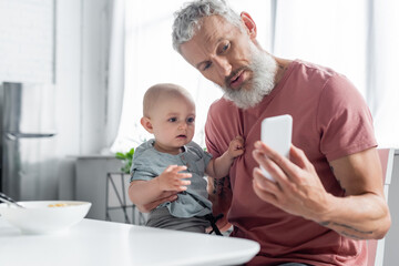Mature father holding smartphone near daughter and blurred breakfast