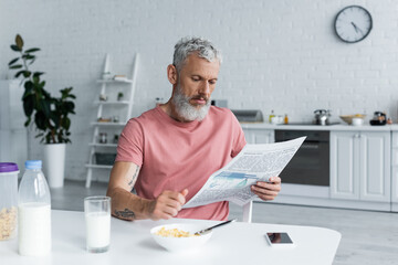 Mature man reading newspaper near corn flakes and smartphone in kitchen