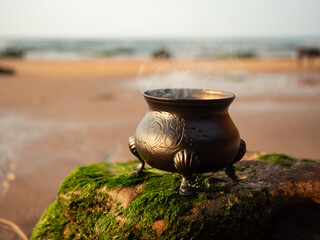 Small black cauldron on a seaweed covered rock on a sandy beach.  Smoke rises as a spell burns within