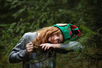 Weinachten, ein  Mädchen mit langen, lockigen Haaren sieht aus wie ein Wichtel. Sie hat eine grüne Wichtelmütze auf. Wichtel steht im Wald.. Sie liegt mit dem Kopf auf dem Tannenzweizweig  und lächelt
