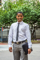 Dressing in white shirt, a black tie, gray pants, carrying a briefcase, a young handsome black college student is standing on a campus, smilingly looking at you..