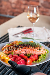 Um pedaço de salmão assado grelhado pimenta limão batata rustica e sal em um prato com folhas de alface e tomate cereja. fundo de tijolos rusticos
e vinha branco
