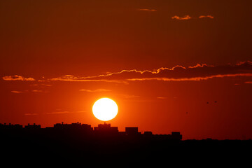Naklejka premium Sunset over city, scenic view. Setting sun and orange sky with dark dramatic clouds above black silhouettes of residential buildings