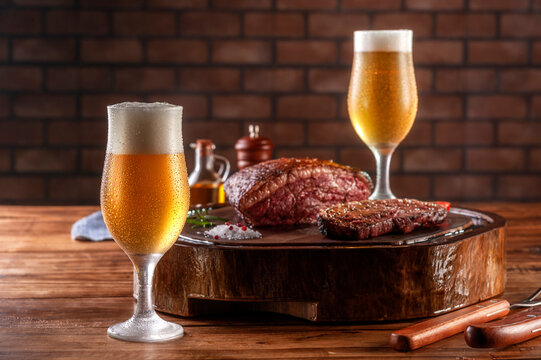 Two sweaty cold tulipa glasses of beer with grilled sliced cap rump steak on wooden cutting board (Brazilian picanha).