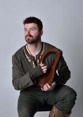 Full length  portrait of  young handsome man  wearing  medieval Celtic adventurer costume playing a small musical harp, isolated on studio background.