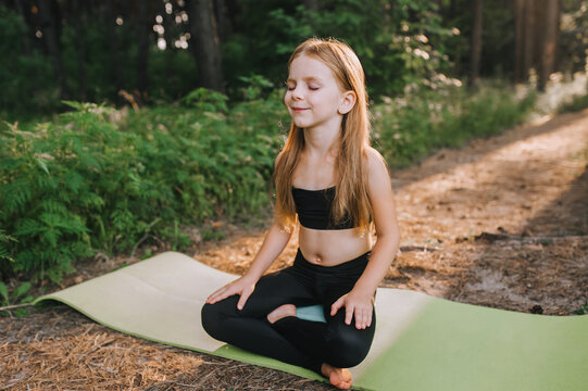 Portrait of a beautiful blonde preschool girl, a professional athlete child in a black suit, sitting on a green rug in a lotus position, crossing her legs, and meditating, training yoga exercises.