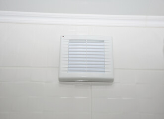 A close-up of a bathroom vent fan, exhaust fan, bathroom ventilation with white grill installed on a white tiled wall.