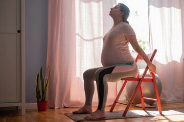 Pregnant woman working out indoors practicing yoga at home using chair