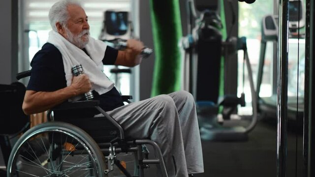 Old cheerful man with a disability exercising with dumbbells in the gym. Person in a wheelchair does arm strengthening exercises