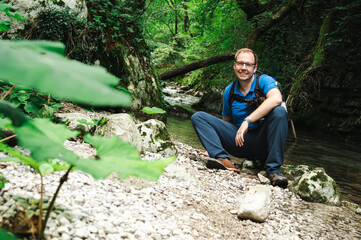 Smiling male photographer with backpack and camera sitting by a stream in the jungle