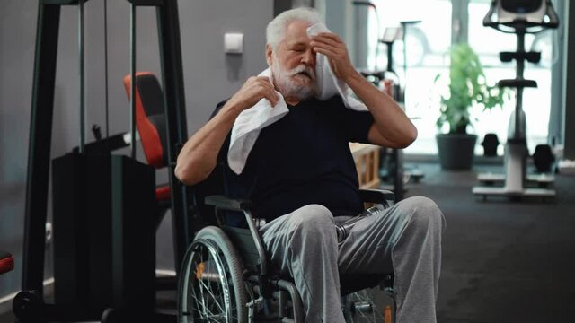 Tired old man in a wheelchair wipes himself with a towel after training in the gym