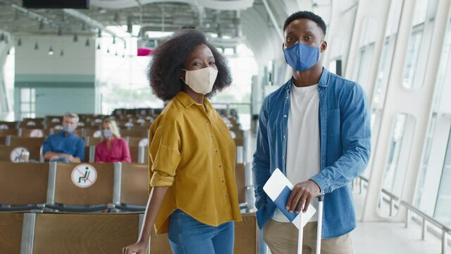 Portrait shot of the young multiracial happy couple wearing protective masks standing in the airport hall and smiling joyfully to the camera