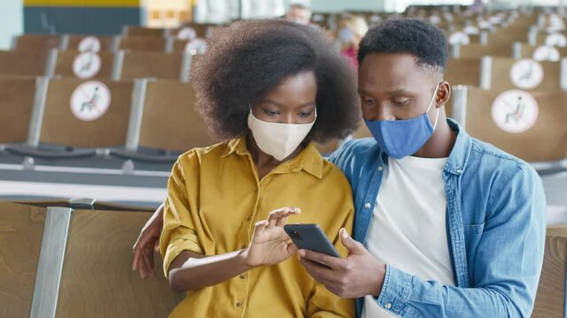Brunette multiracial young beautiful woman showing something to her boyfriend on the smartphone while they waiting for the flight. People wearing protective masks