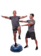 An elderly man doing a balance exercise, on one leg, on a hemisphere ball. With help of a fitness...