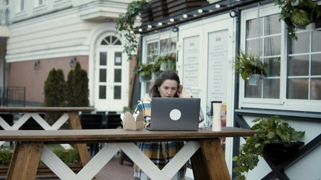 Thoughtful young female entrepreneur reading business information on laptop while working on project at table of outdoor cafe in autumn day