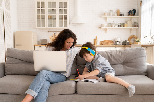 Work from home with kid: curious loving mom look at small son in headphones drawing, sit with laptop on couch, surf internet for work. Mother office worker or freelancer on lockdown. Stay home concept