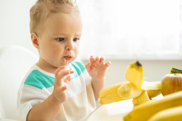 funny baby boy sitting at the table in child chair eating banana on white kitchen.