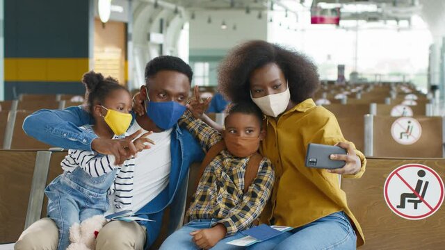 Young afro american spouses wearing protective masks sitting in the airport and taking selfie photos with their kids to the camera of smartphone