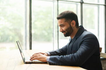 Fototapeta premium Cheeful Indian businessman using laptop computer for remote work, happy young bearded male ceo manager in formal suit typing on the keyboard sitting at the desk in loft office