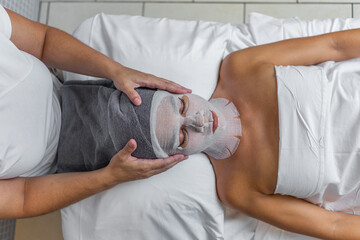 Beautician applying a towel on head of a woman who is covered her face with gauze for skin treatment