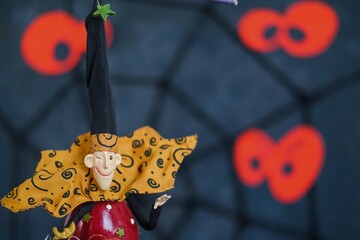 Halloween party background composition, invitation card or club flyer with copy space. Focus on head of colorful freak figurine on dark blurred background of cobwebs and defocused jack lanterns.