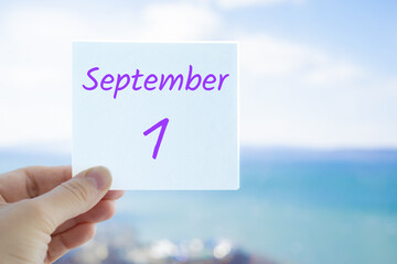 1st September. First day of autumn. Knowledge day. Hand holding sticker with text September 1 on the blurred background of the sea and sky. Copy space for text. Month in calendar concept