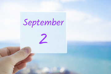 2nd September. Hand holding sticker with text September 2 on the blurred background of the sea and sky. Copy space for text. Month in calendar concept