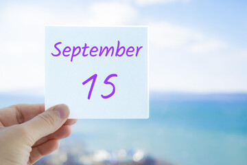 15th September. Hand holding sticker with text September 15 on the blurred background of the sea and sky. Copy space for text. Month in calendar concept