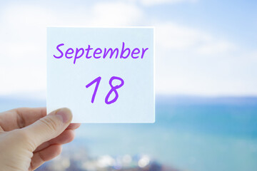 18th September. Hand holding sticker with text September 18 on the blurred background of the sea and sky. Copy space for text. Month in calendar concept