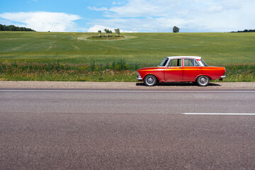 Red retro car is standing on the side of the road near an endless green field. Car trip among breathtaking beauties of nature