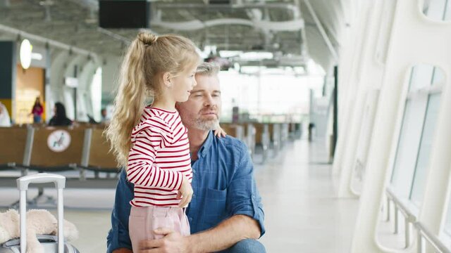 Senior handsome caucasian father showing to his daughter airplanes through the airport window. Little pretty girl waiting for departure or arrival with father. Traveling, tourism, weekends concept