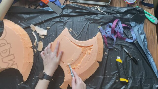 Hands cut hatchet form from foam polystyrene with stationery knife on garbage bag. Craftsman's workshop. Woman makes viking axe for cosplay festival. Diy celtic weapon on wooden table.