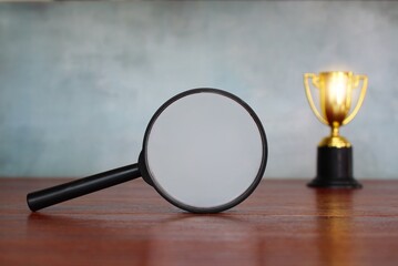 Selective focus image of magnifying glass and shining gold cup trophy on wooden table