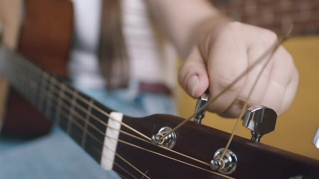 Womans hand tunes an acoustic guitar by pulling string by twisting guitar pegs, closeup. Musician tunes his instrument before performance
